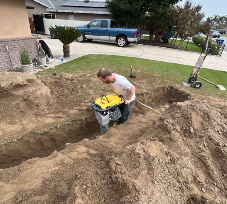 A focused PLUMBER IN WASCO, CA using a high-powered soil compactor within a deep trench, ensuring a stable foundation and proper backfilling for a newly installed residential main line.